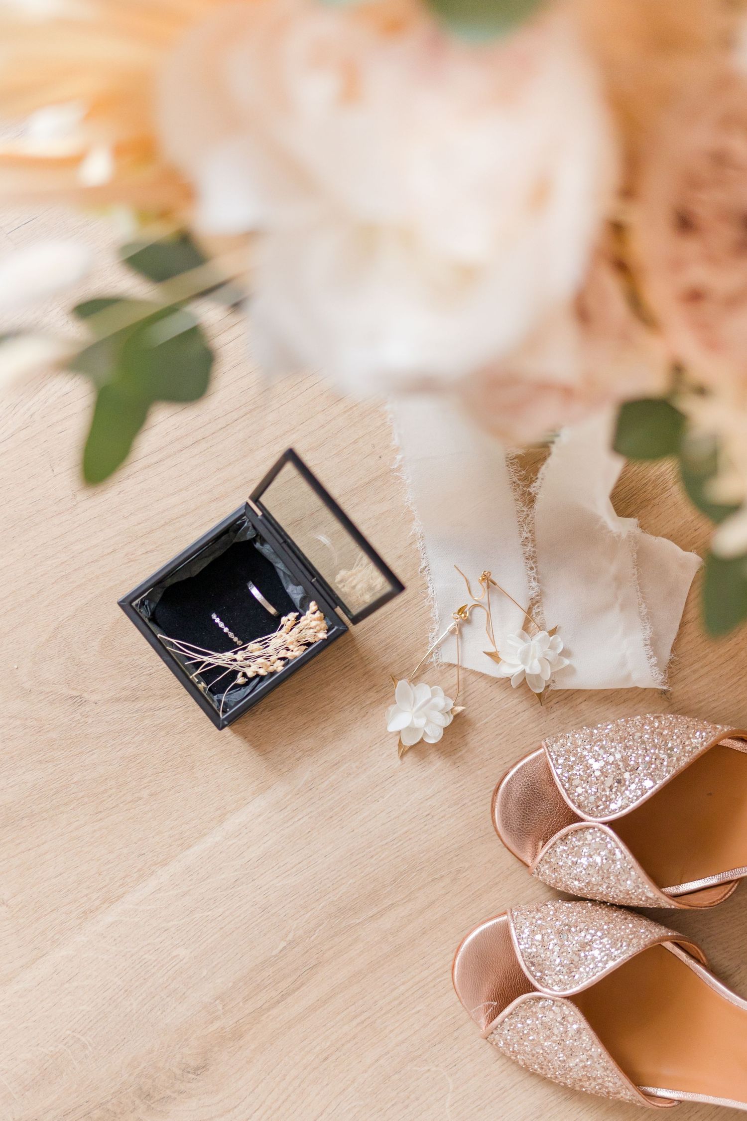 Sparkly bridal shoes and a small black box arranged on a beige surface with delicate floral elements.
