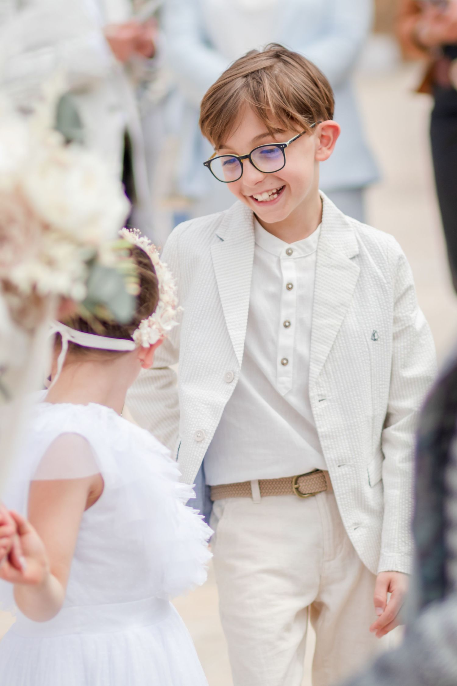 Young children in white formal wear smile and interact at wedding celebration.