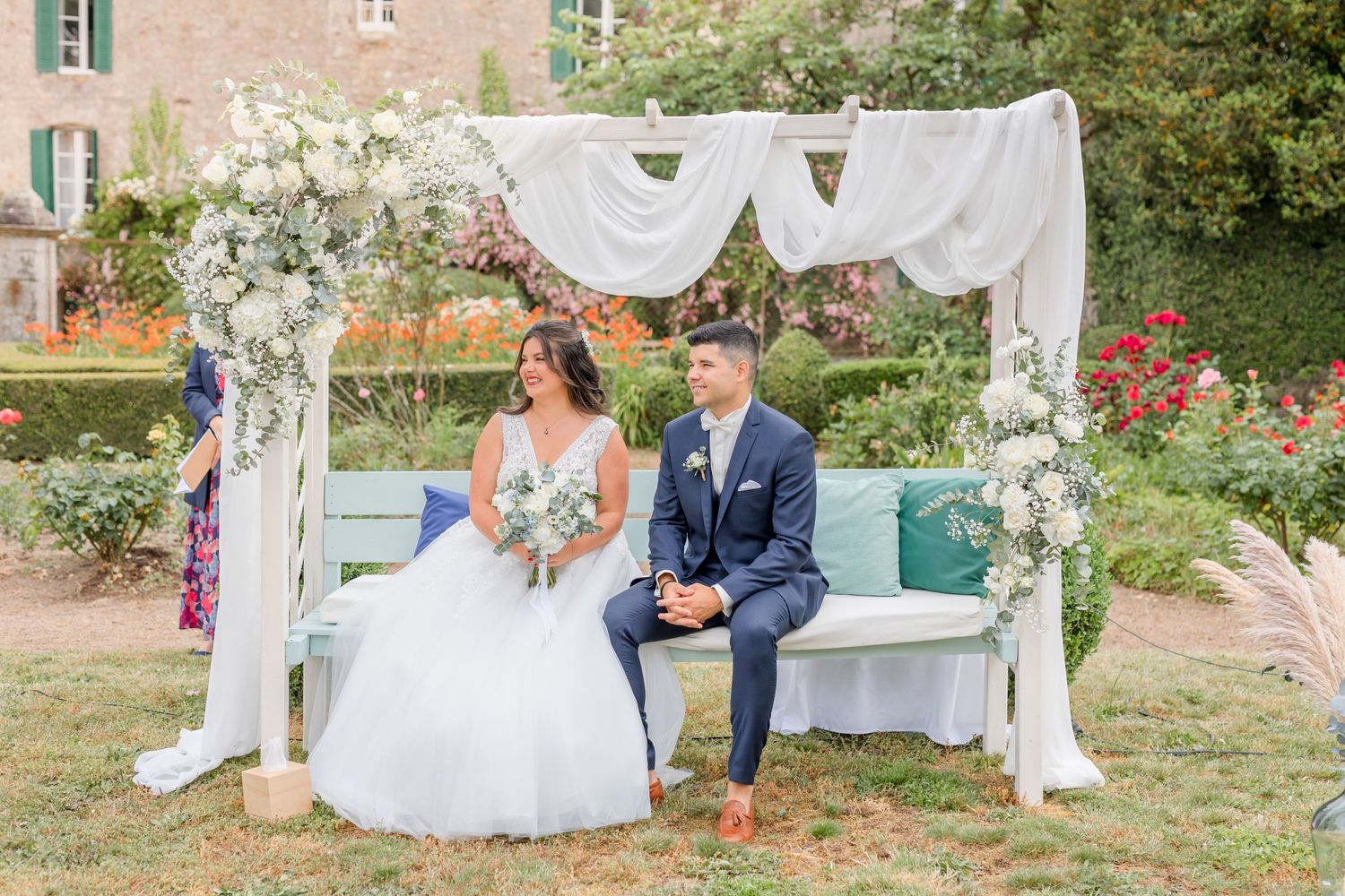 Couple seated under decorated white arch at romantic garden wedding.