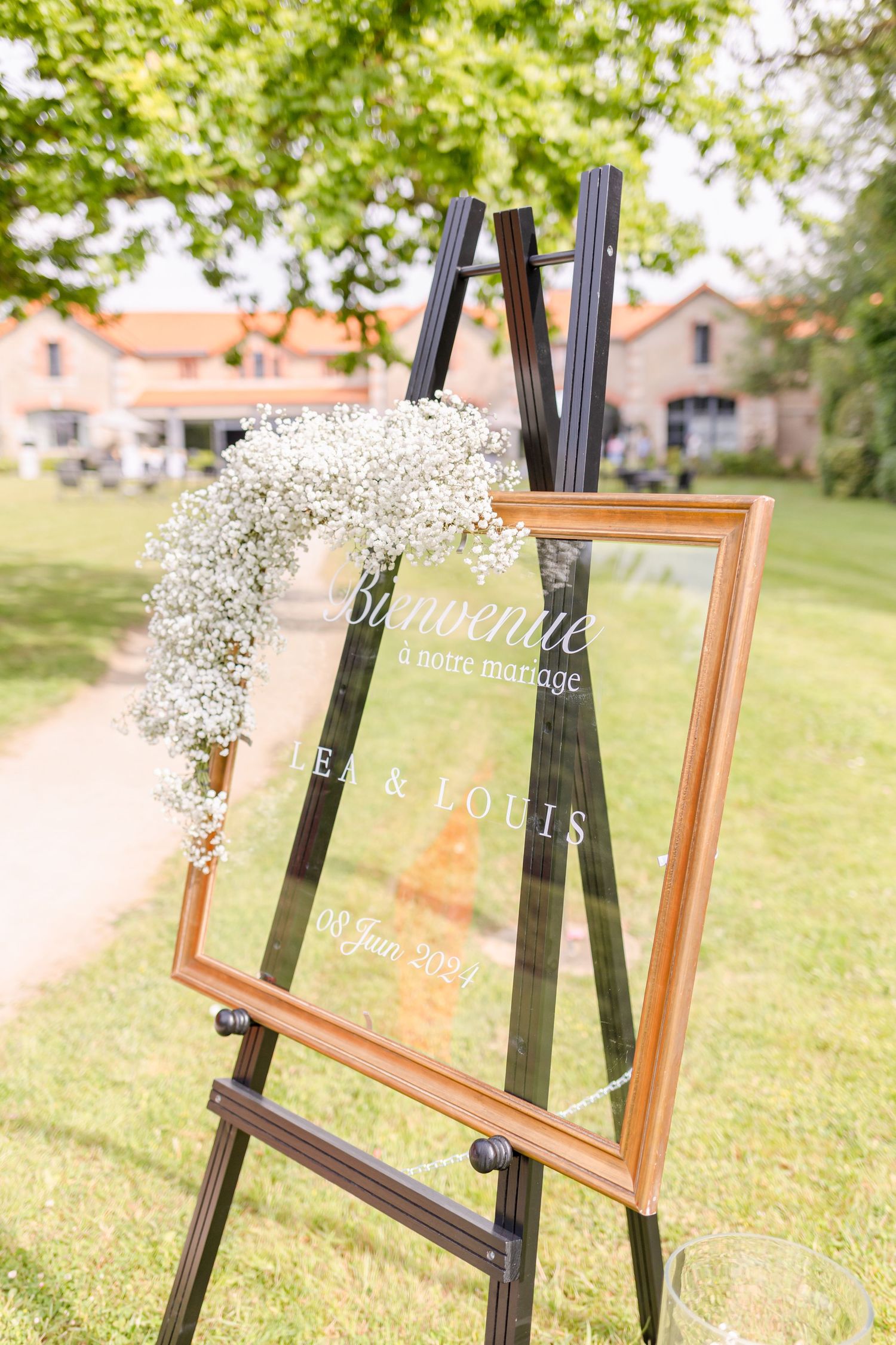 Wedding welcome sign on an easel decorated with baby's breath flowers on a grassy lawn with buildings in background.