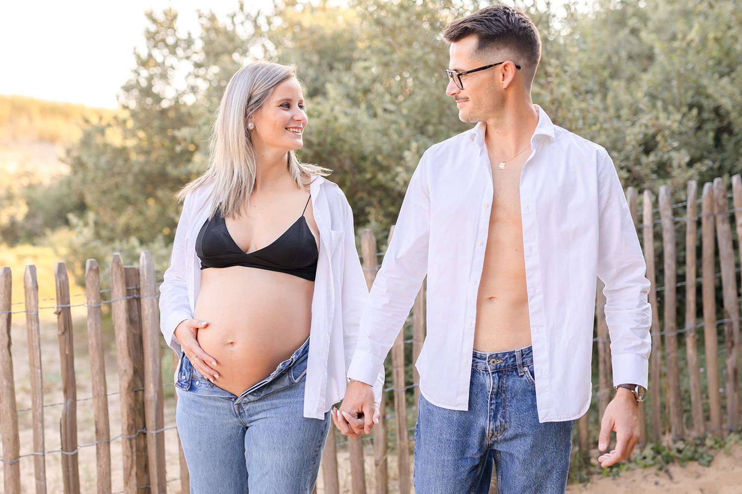 Intimate outdoor photoshoot shows couple in casual white shirt and denim outfits against wooden fence at sunset.