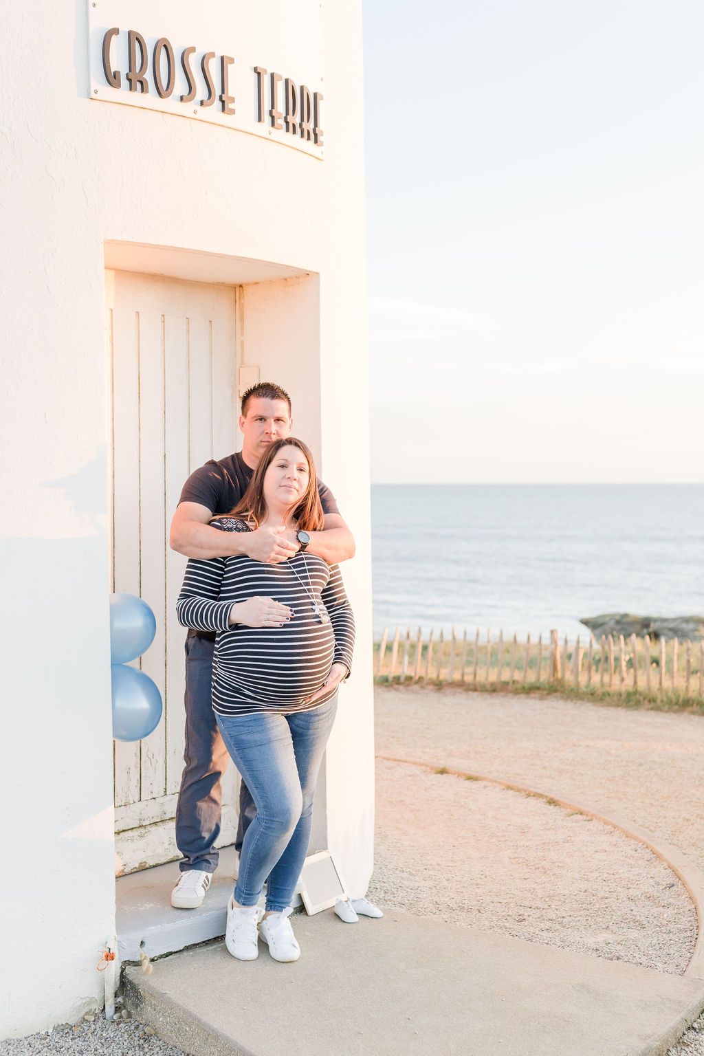 Couple embracing by lighthouse entrance during seaside maternity session.