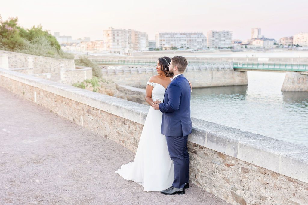 Wedding couple stands together on a stone bridge with waterfront city views in the background.