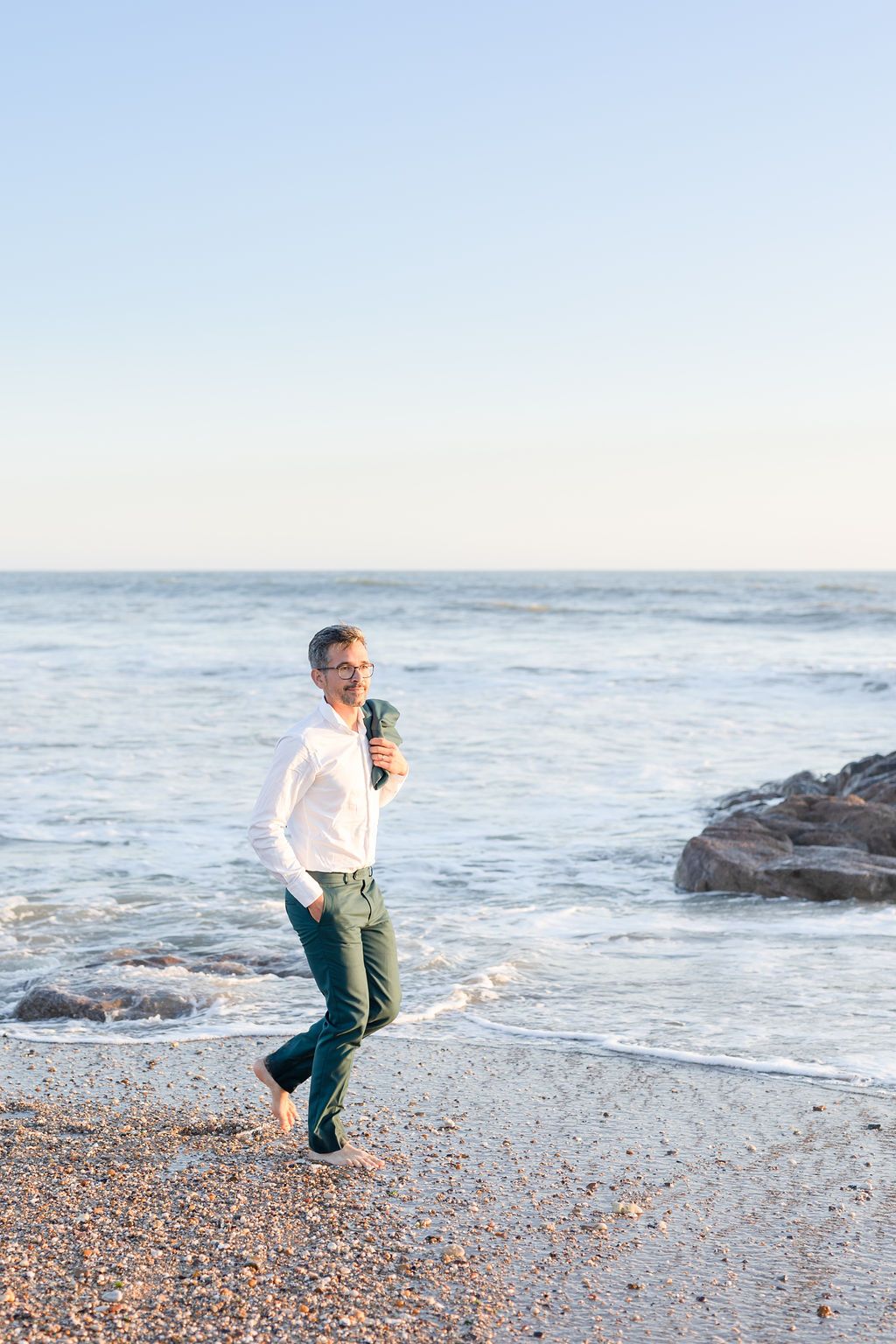 A person in casual attire walks barefoot along a rocky beach shore at sunset.