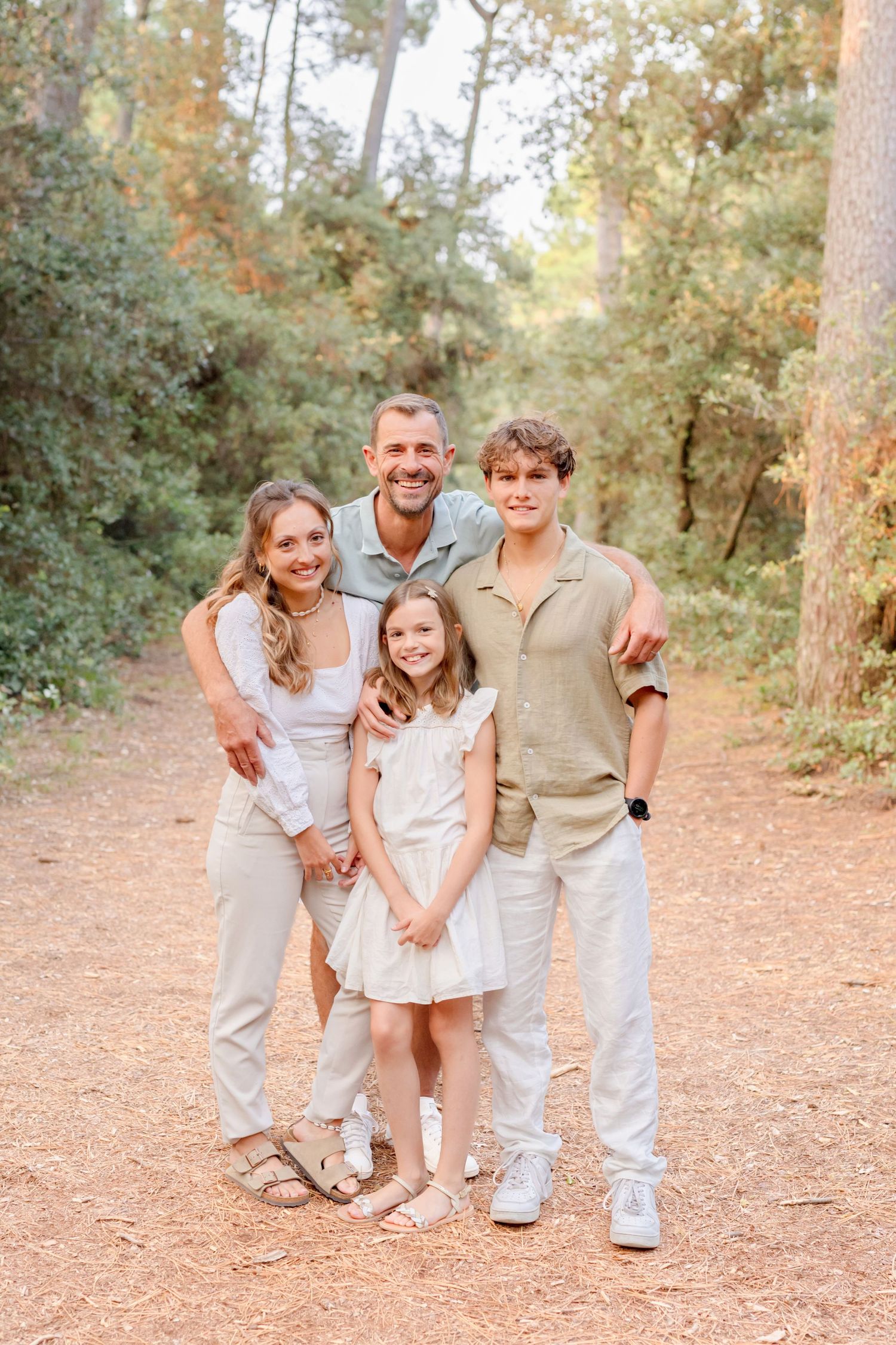 A family wearing coordinated light-colored outfits poses together on a wooded trail.
