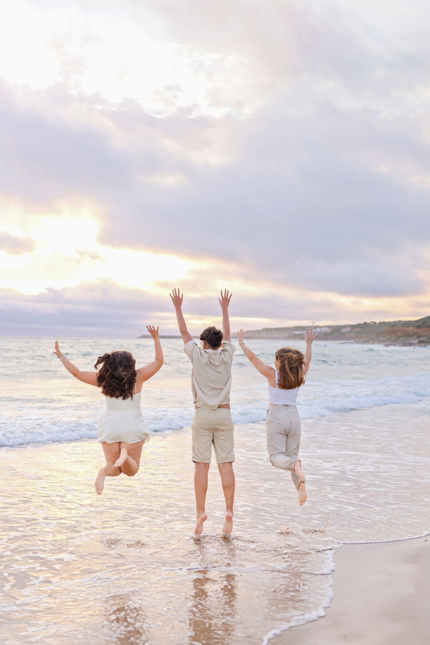 Three friends jump with raised arms on a beach at sunset creating a joyful silhouette.