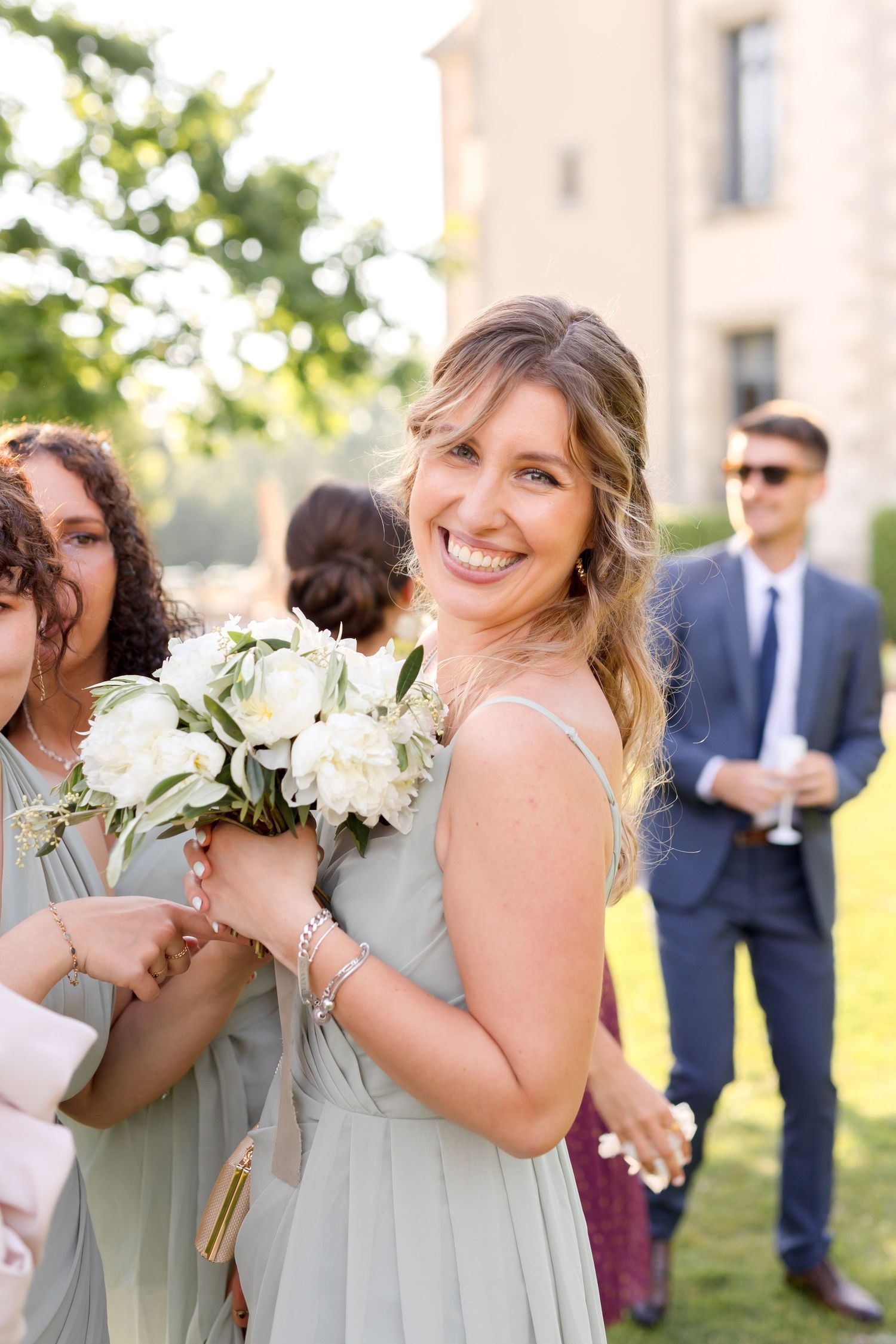 Bridesmaids in sage green dresses hold white floral bouquets while celebrating outdoors.