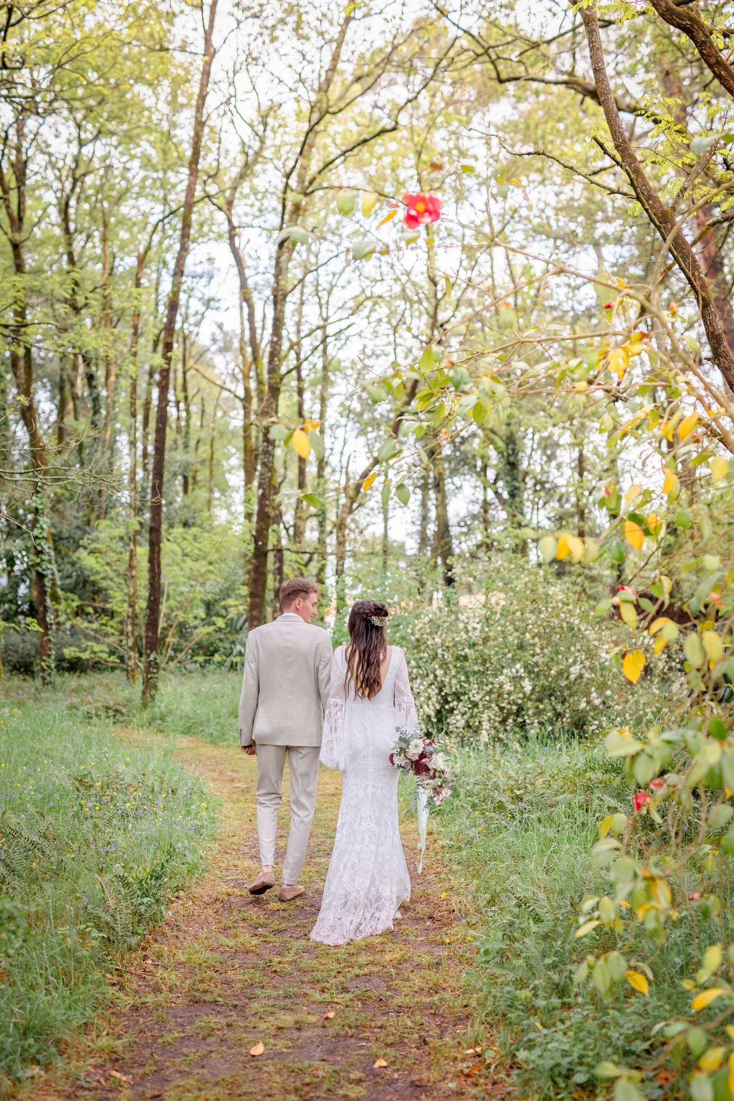 A couple walking down a wooded path surrounded by autumn trees and fallen leaves.