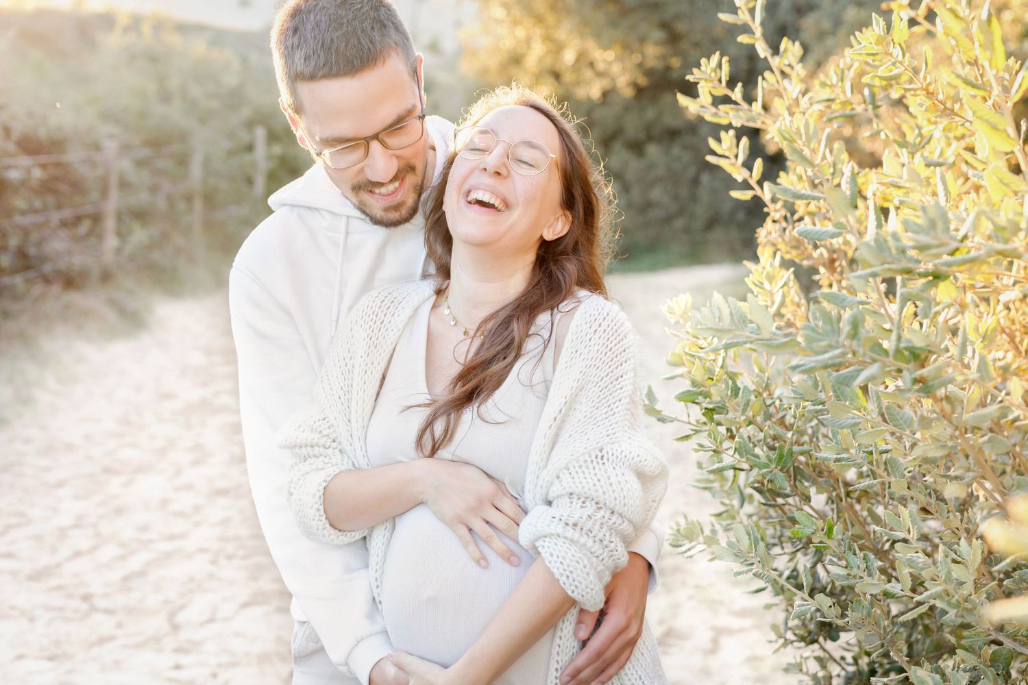 Couple in white outfits share joyful embrace and laughter during outdoor sunset photoshoot.