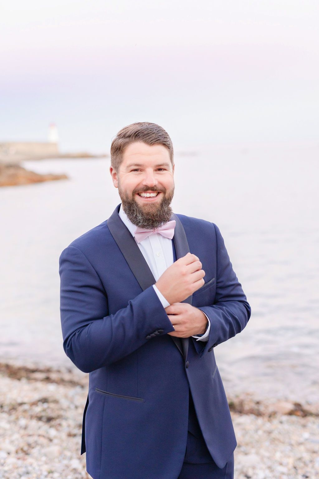 A happy groom in a navy suit adjusts his cufflink by the seaside.