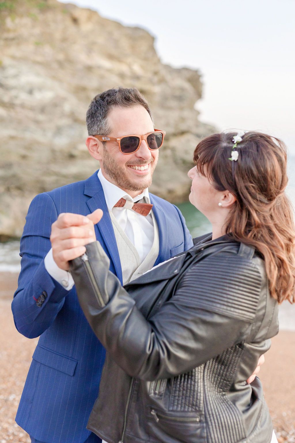 A playful wedding couple dances on the beach wearing formal attire and casual leather jacket.