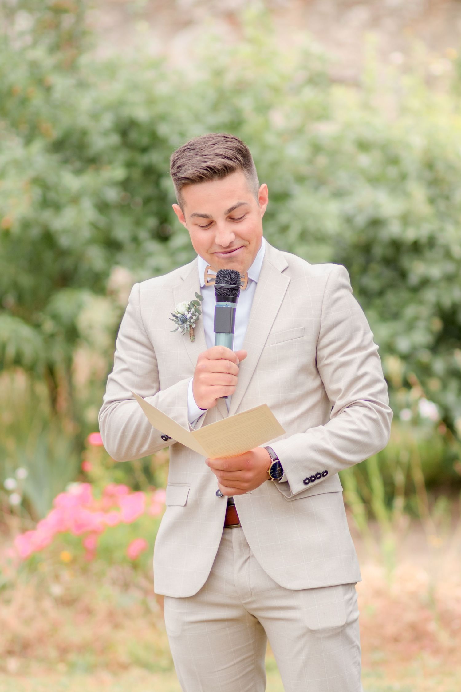 Groom in beige suit reading from papers during outdoor wedding ceremony.