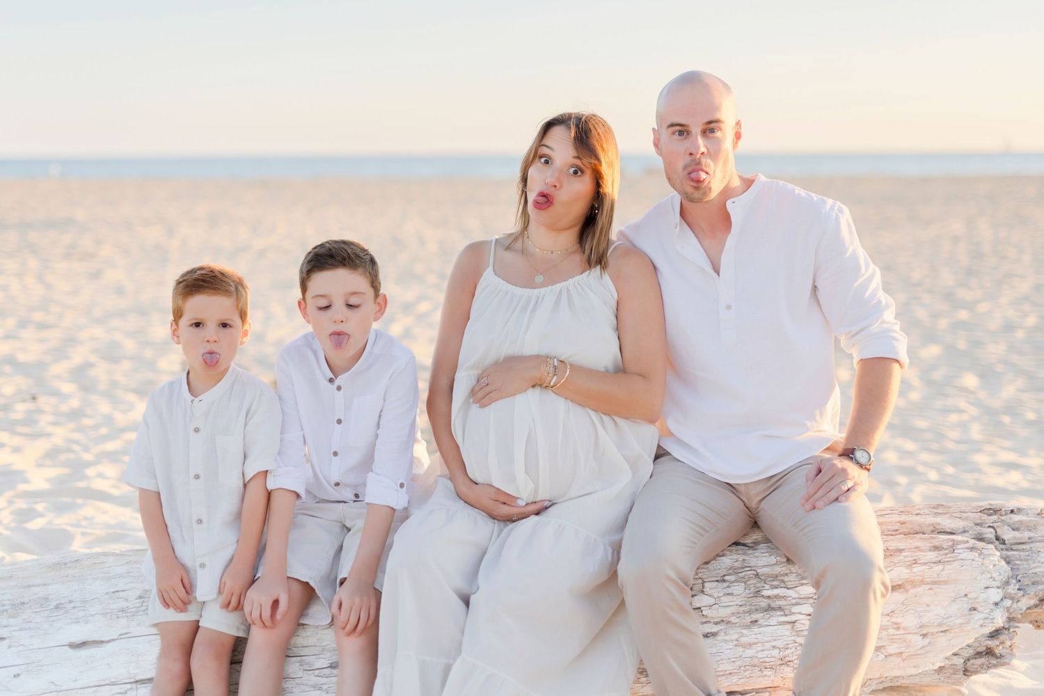 Family of four wearing coordinated white outfits poses together on beach at sunset.