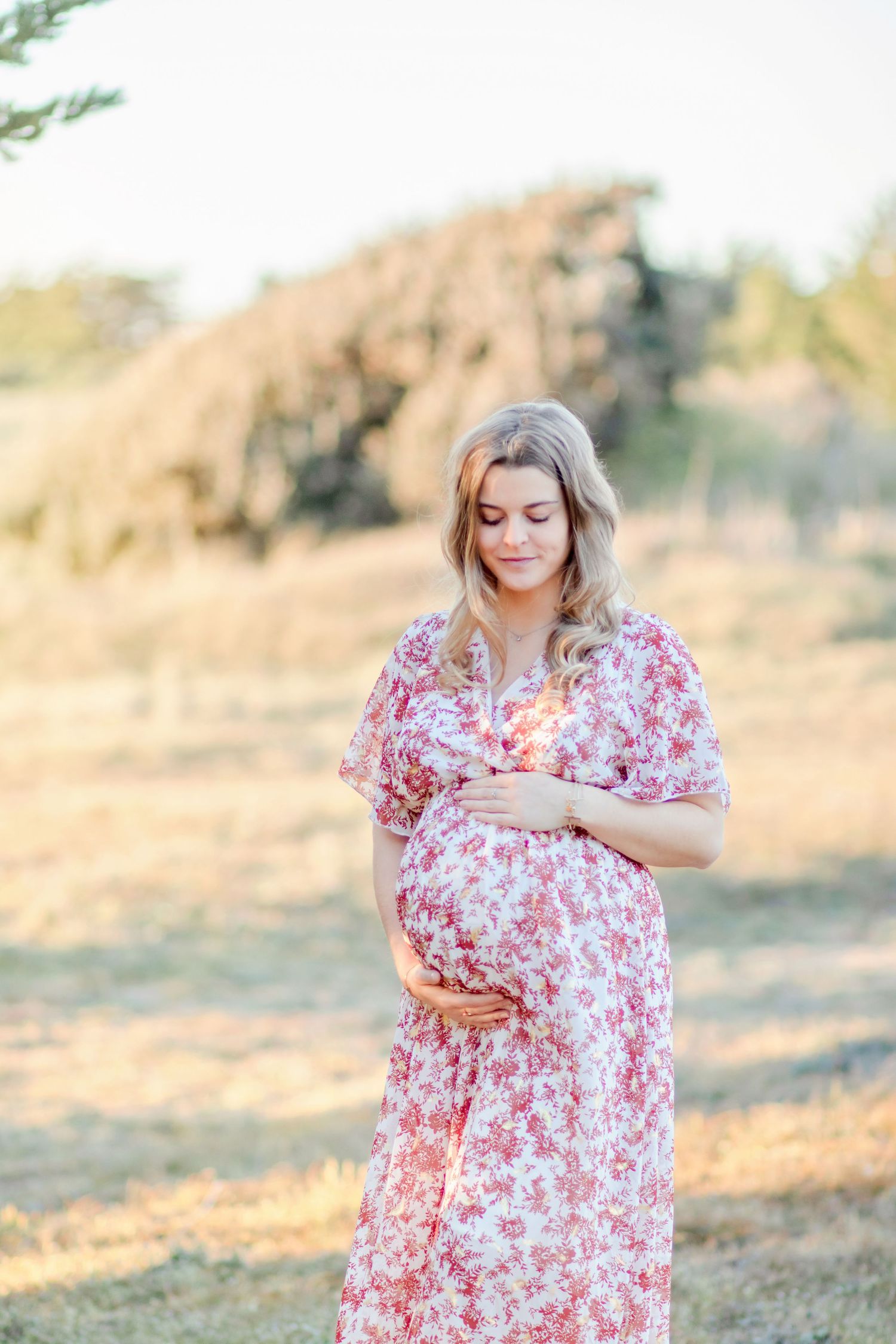 Woman in pink floral dress cradles baby bump in outdoor setting.