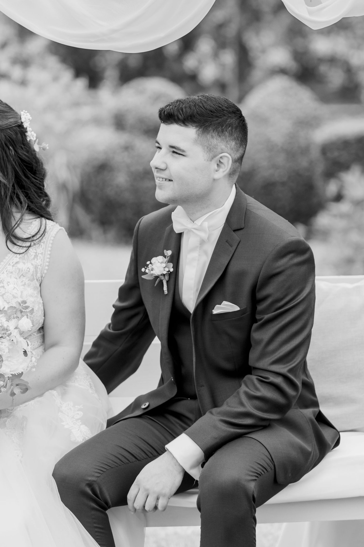Groom in formal black suit sitting during outdoor wedding ceremony.