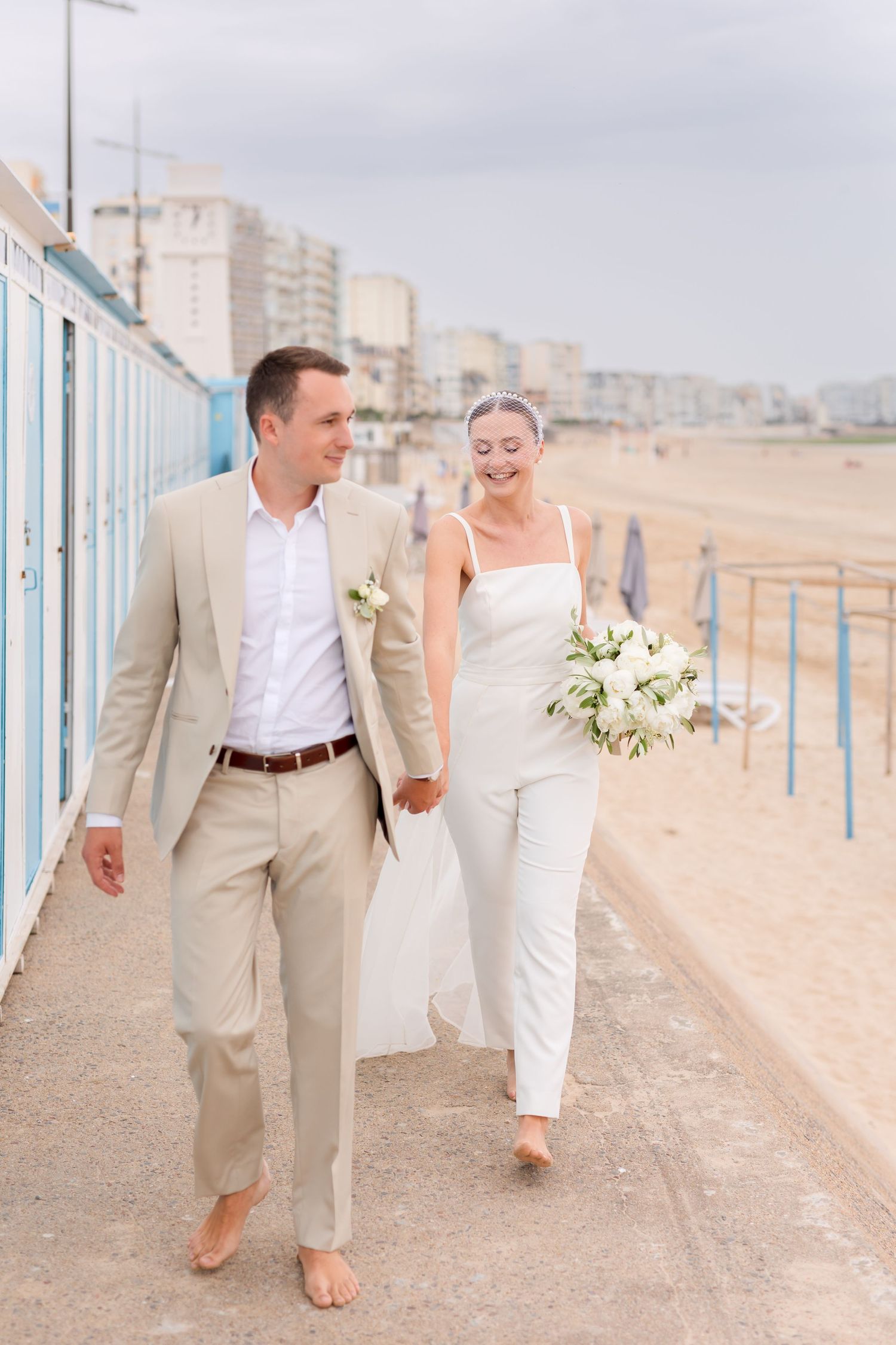 A barefoot couple strolls along a beach promenade with blue beach huts in the background.