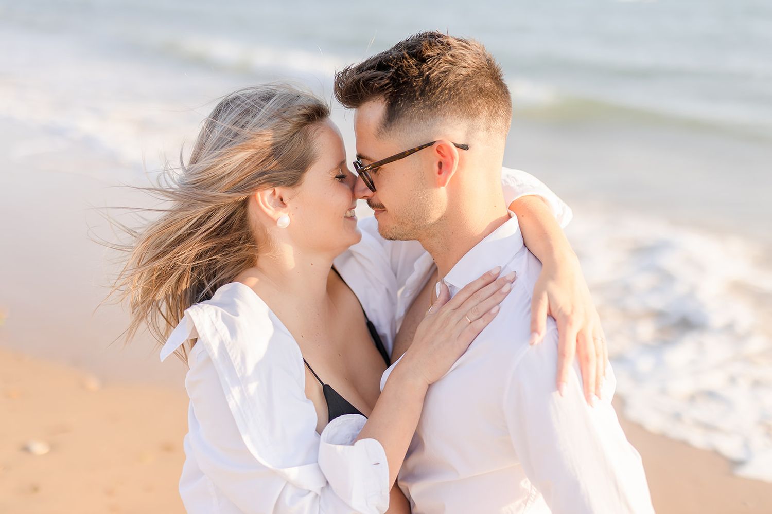 A romantic couple in white shirts embrace on a sunny beach with ocean waves in the background.