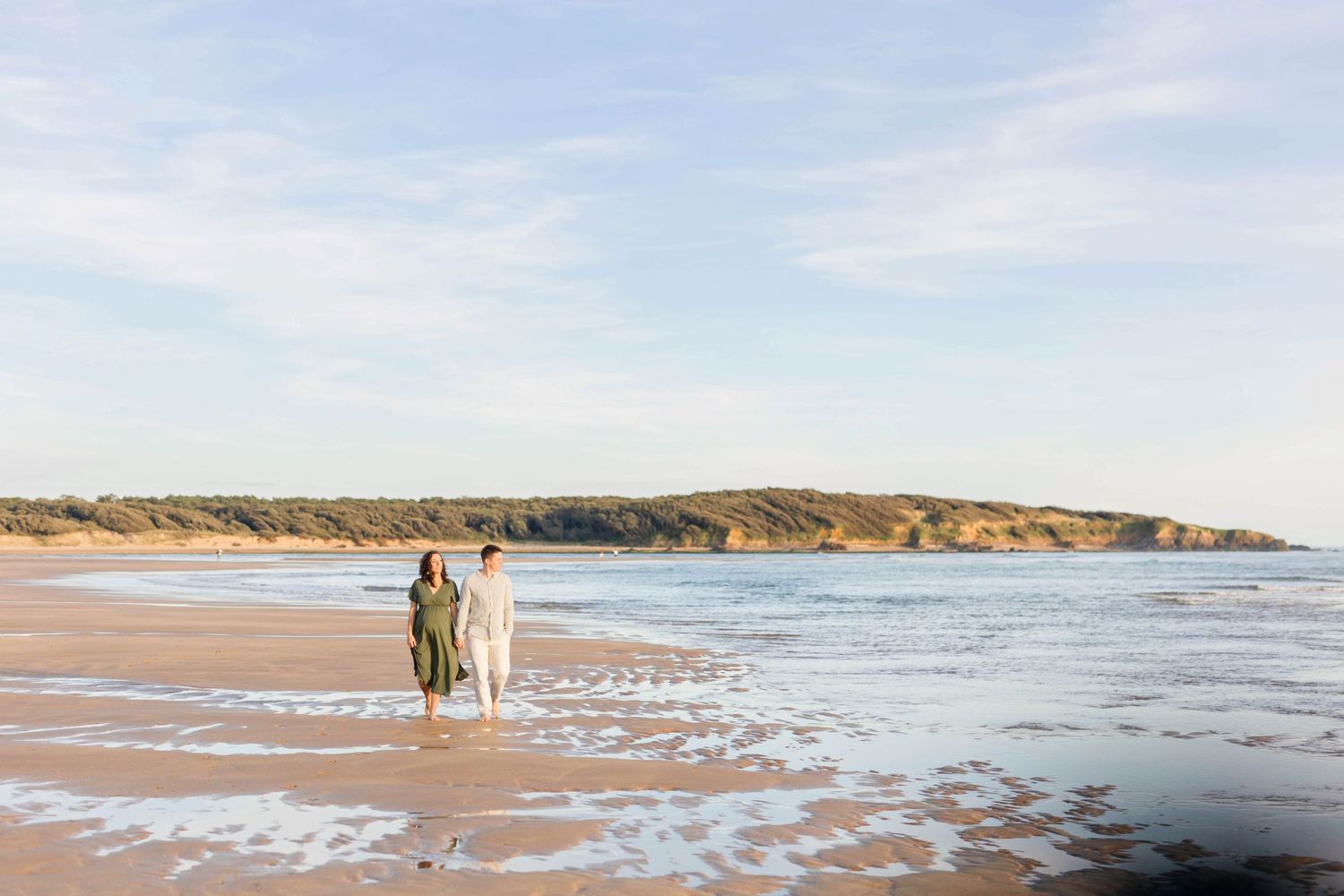 A couple stands at water's edge on expansive beach with coastal cliffs in background.