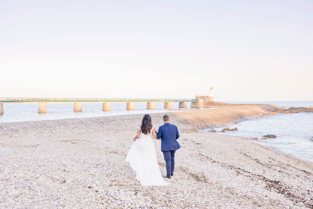 Beach wedding couple walking along rocky coastal path with lighthouse in the distance.