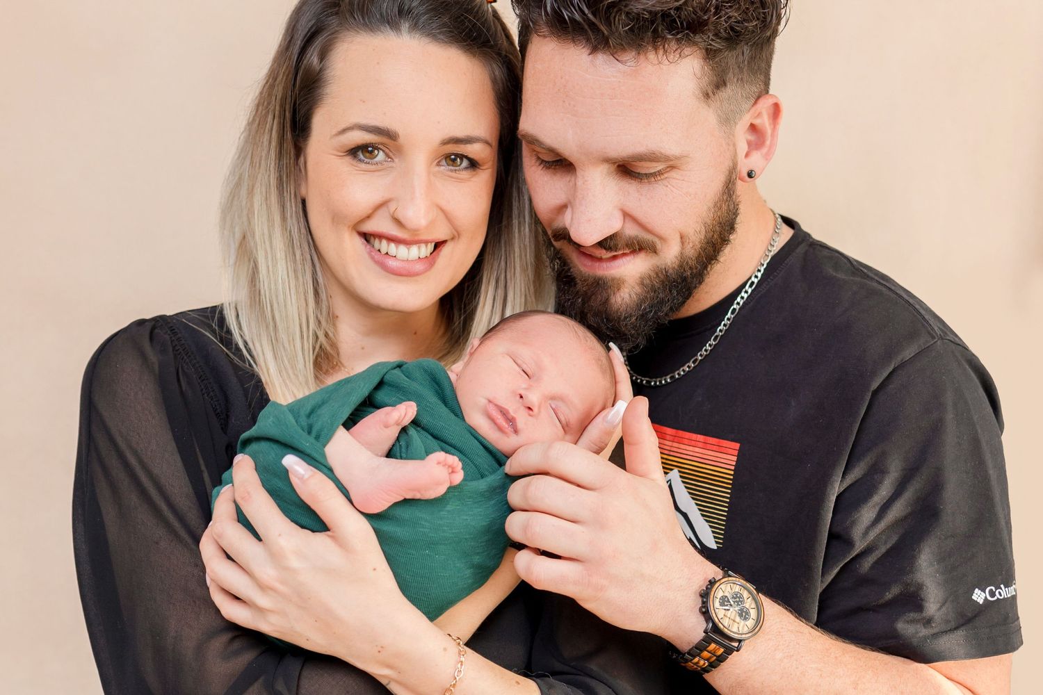 New parents lovingly hold their sleeping newborn baby wrapped in a green swaddle.