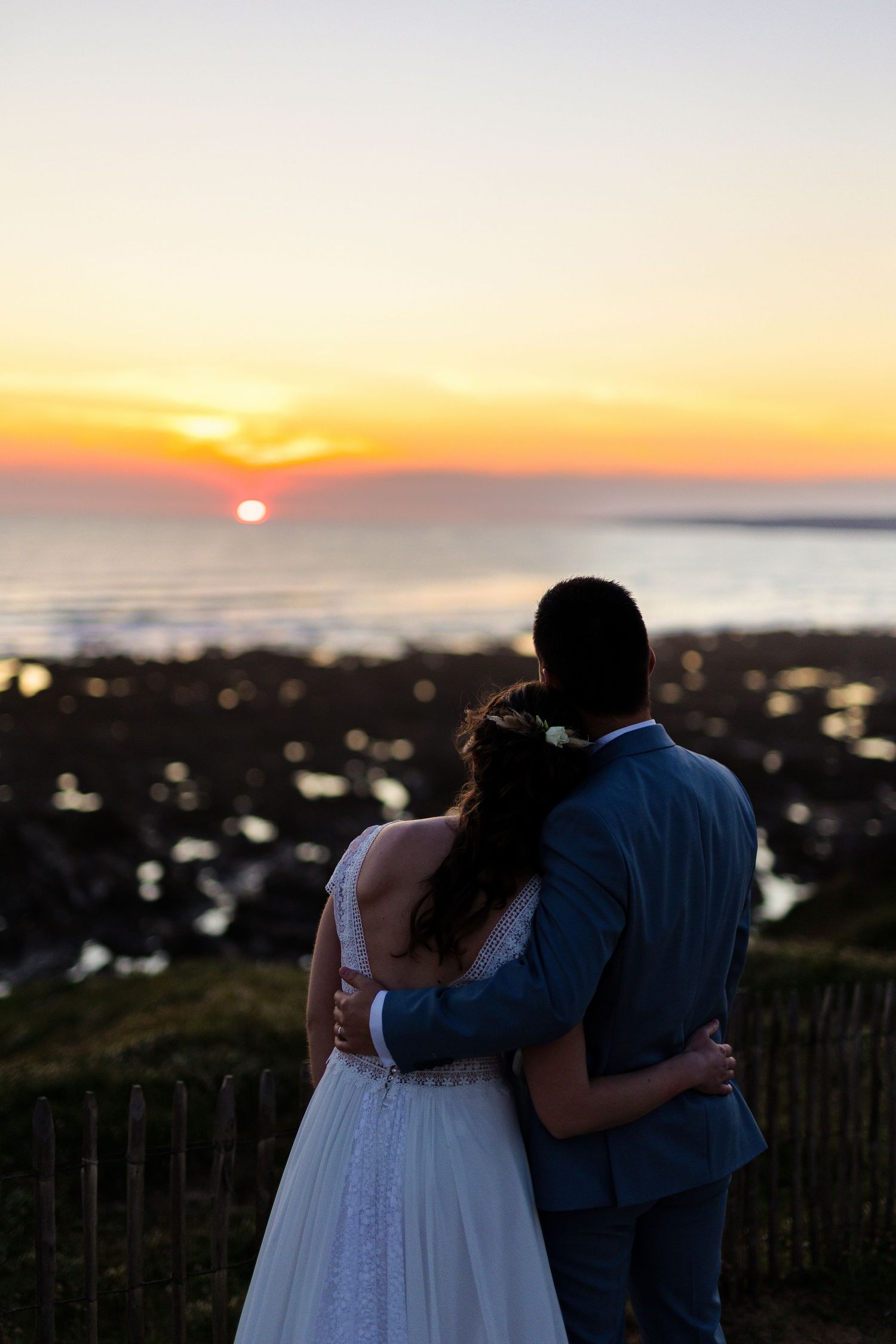 Silhouetted couple watching sunset over ocean from coastal viewpoint.