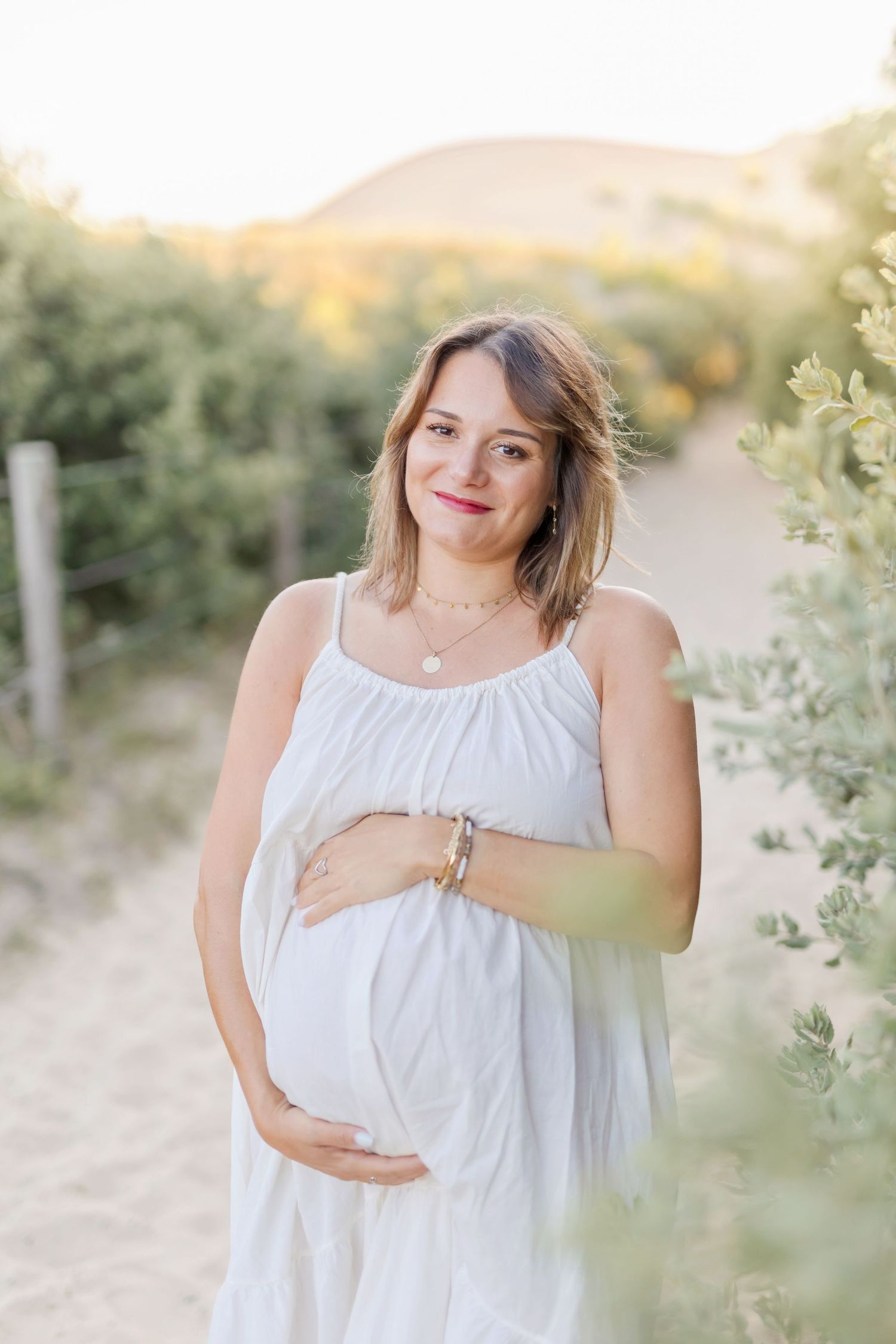 Glowing maternity portrait of woman in white dress standing on nature path at sunset.
