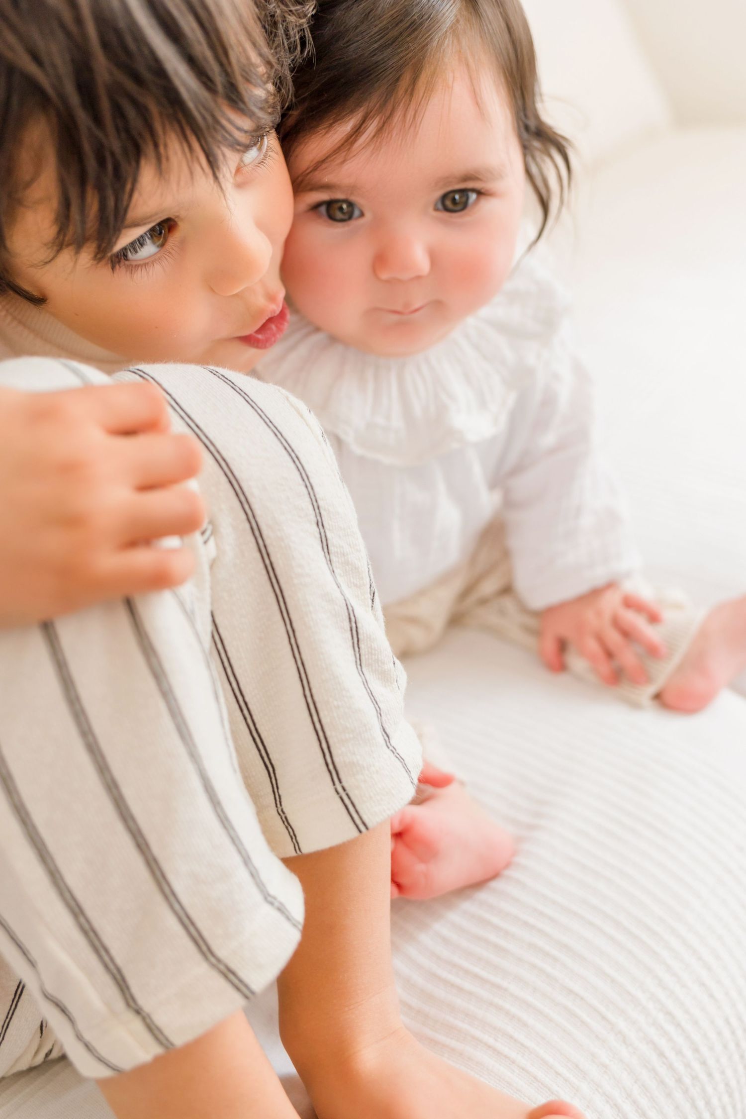 An infant in white clothing lies on cream colored bedding while being kissed.