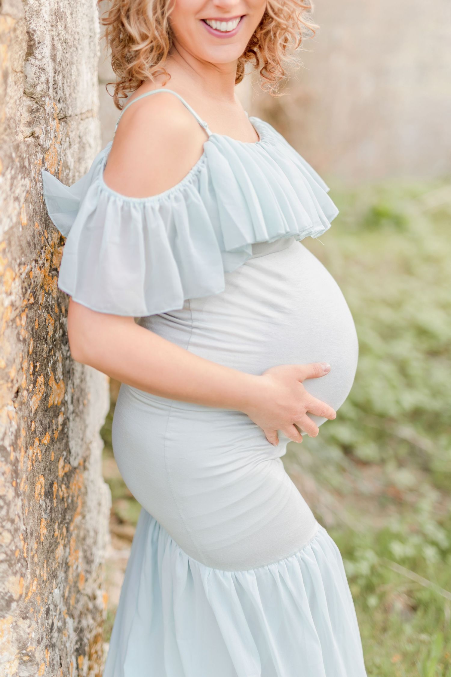 Side view of pregnant woman in pale blue ruffled dress against stone wall.