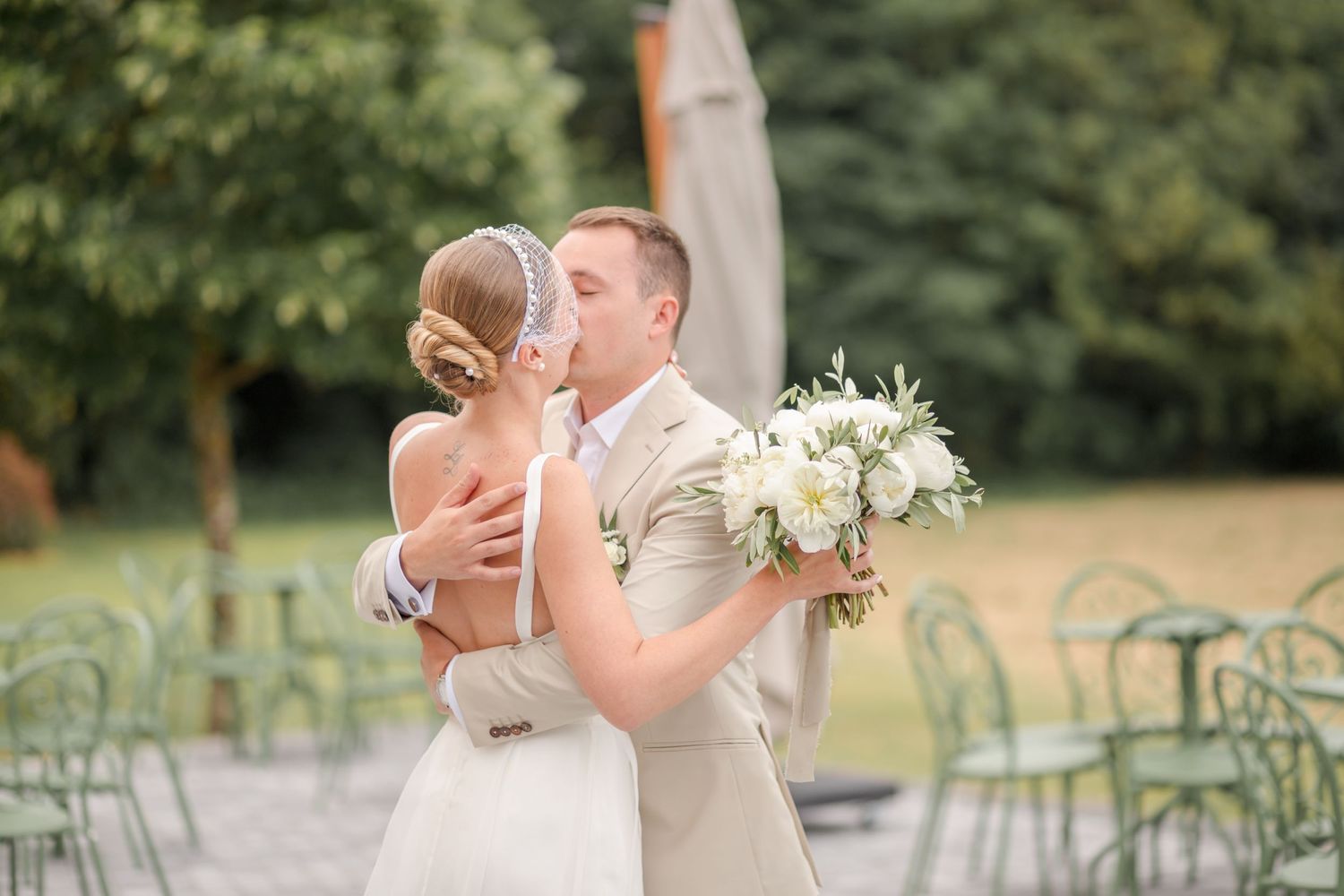 Romantic wedding couple embracing outdoors in an elegant garden setting with cafe chairs.