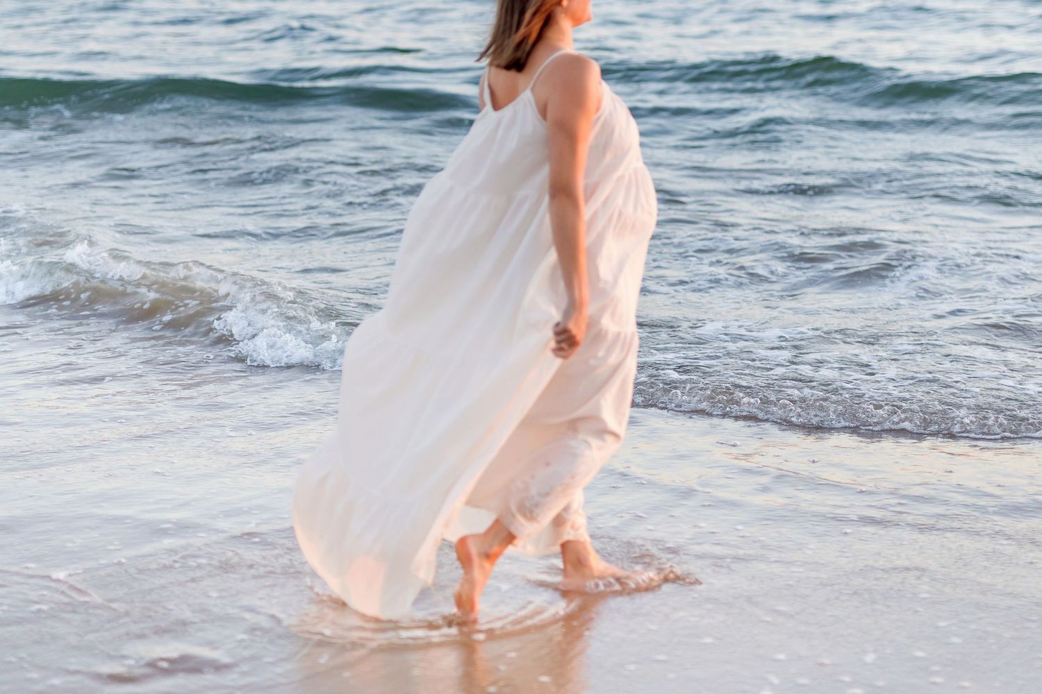 A flowing white dress catches the breeze along a scenic beach shoreline at sunset.