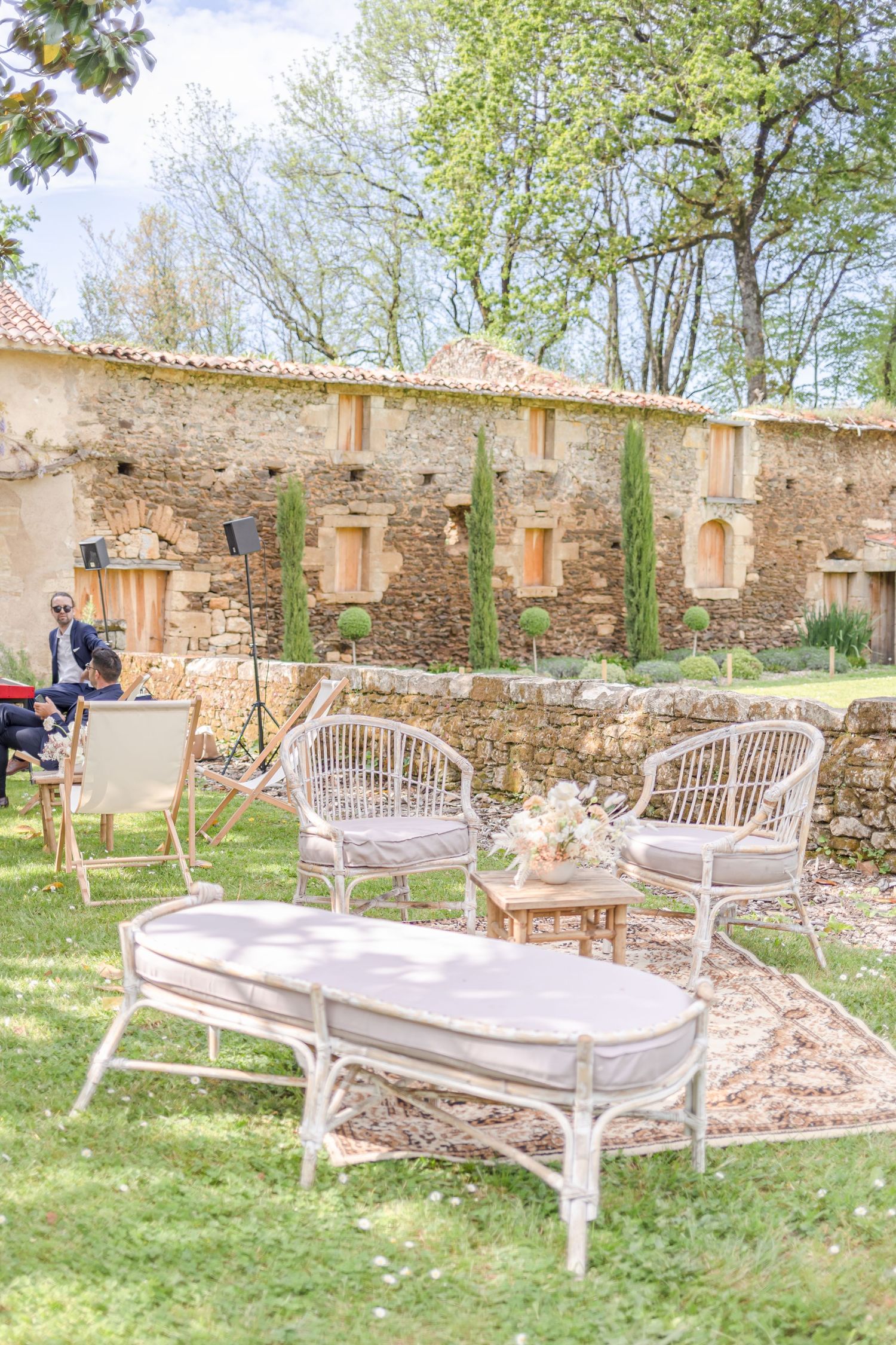 White wicker outdoor furniture arranged in a rustic courtyard setting with stone walls and cypress trees.