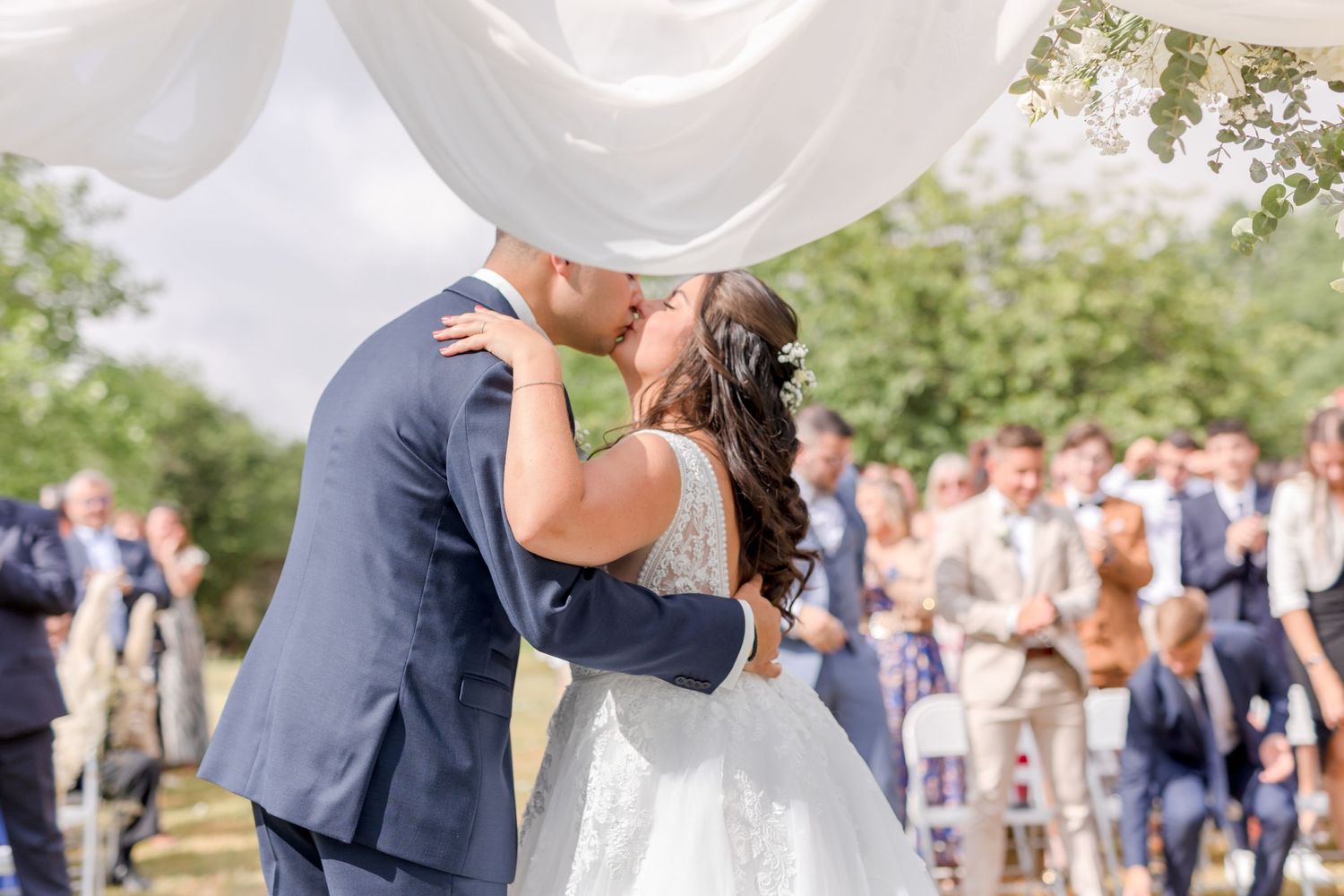 Wedding celebration with guests gathered outdoors under white tent canopy.