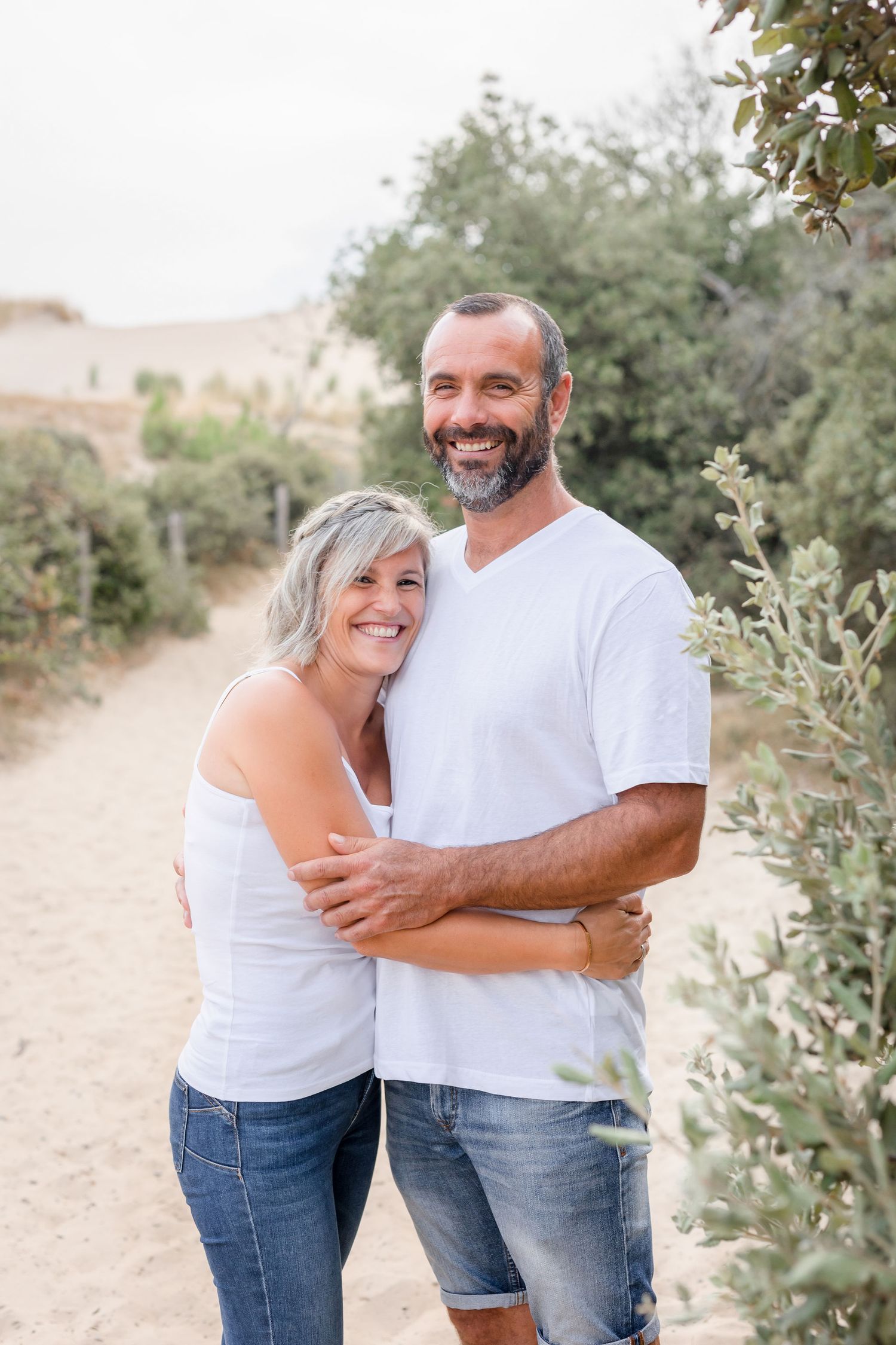 A couple wearing matching white tops and jeans embraces and laughs together in a sunny outdoor setting.