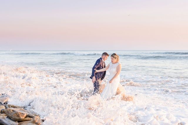 A couple laughs as ocean waves splash around them during their beach wedding photoshoot.