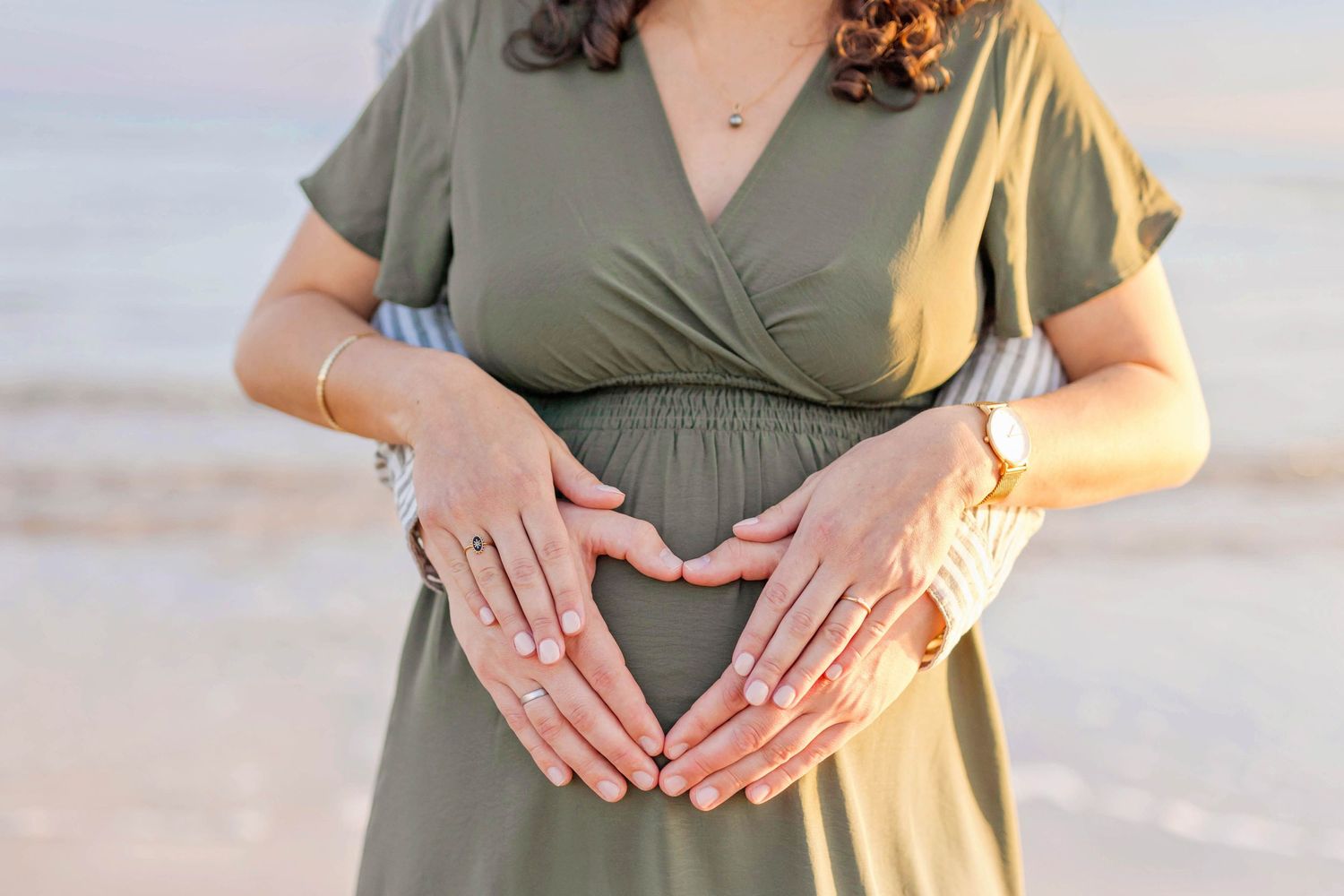 A pregnant woman in a sage green dress holds her baby bump tenderly at the beach during sunset.