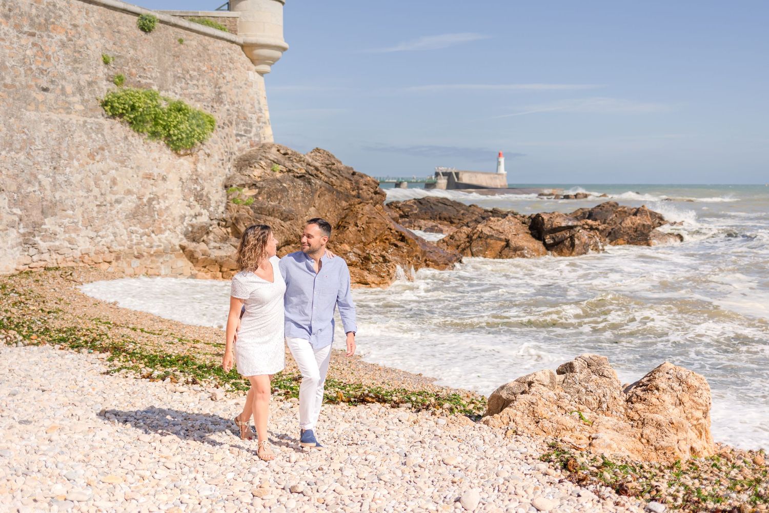 Couple in light summer clothing walking along rocky Mediterranean coastline with lighthouse in distance.