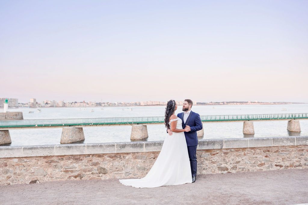 Newlyweds pose along a historic stone wall with a serene waterfront vista in the background.