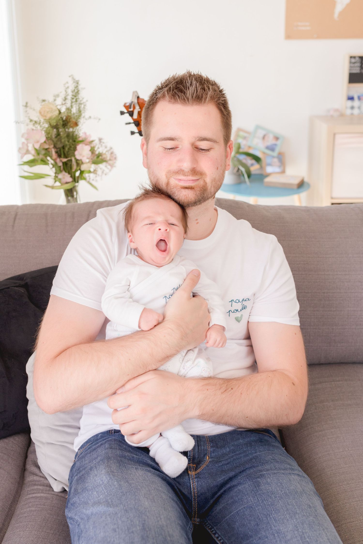 Parent cradling sleeping infant on gray couch in bright living room with flowers.