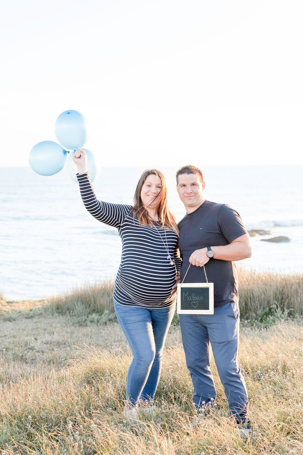 Happy couple celebrating pregnancy announcement with blue balloons and frame by the ocean.