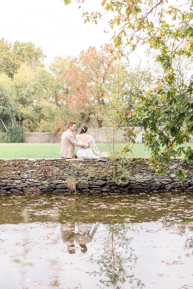 Romantic wedding photos by stone wall with autumn foliage and water reflection.