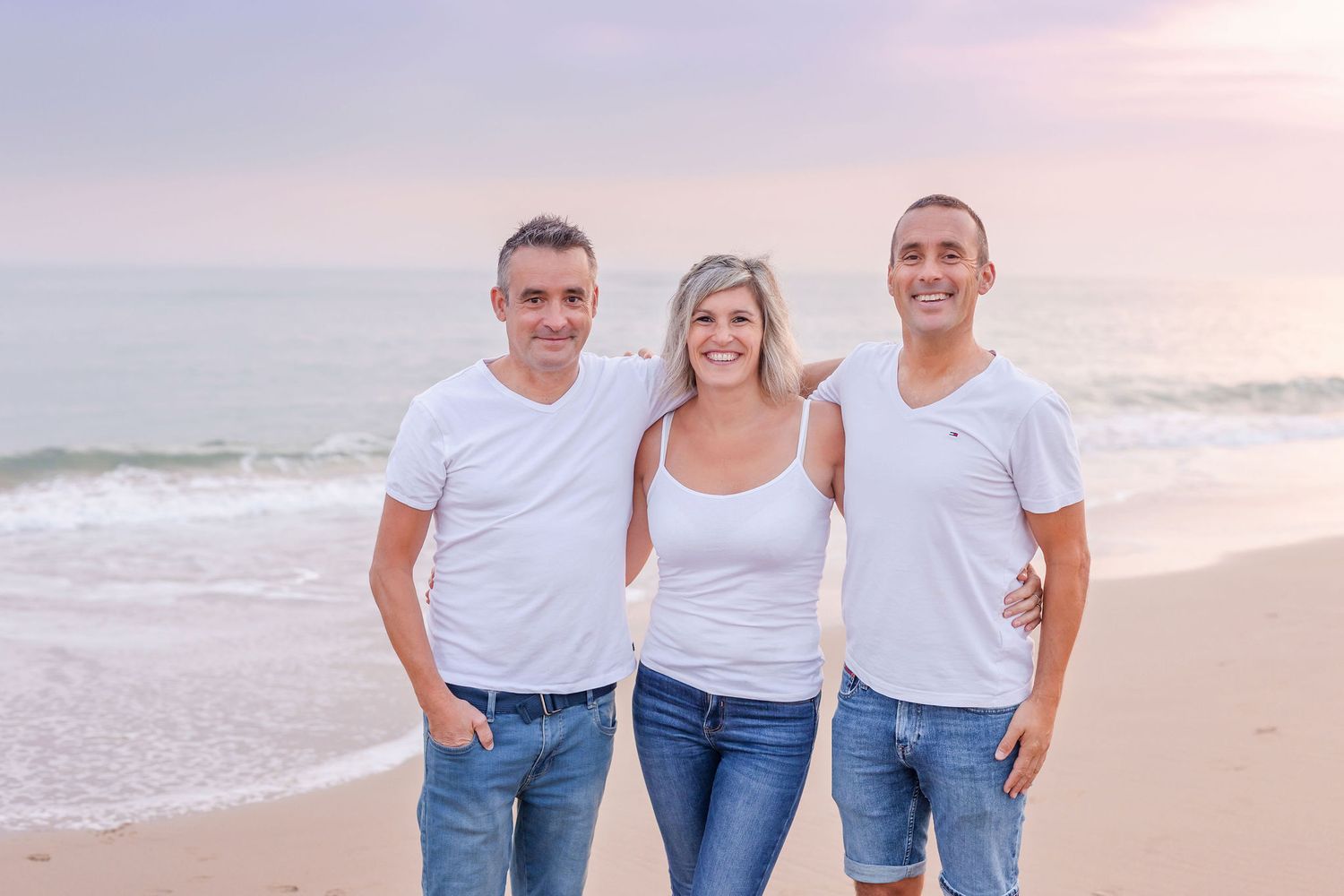 Three people wearing white tops and denim stand together smiling on a beach at sunset.