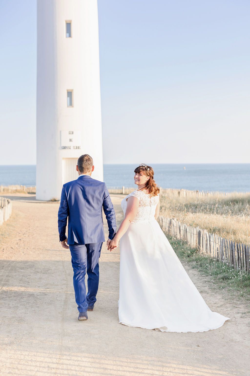A scenic wedding photo showing a couple walking towards a white lighthouse beside the ocean.