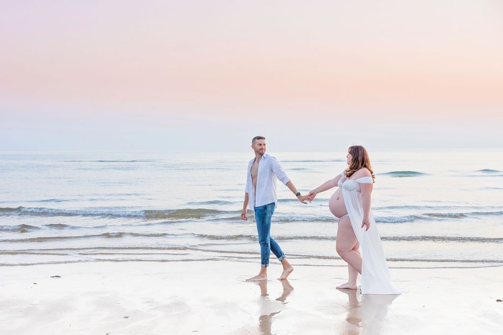 Two people in white clothing dance and play on the beach at sunset with waves rolling in.