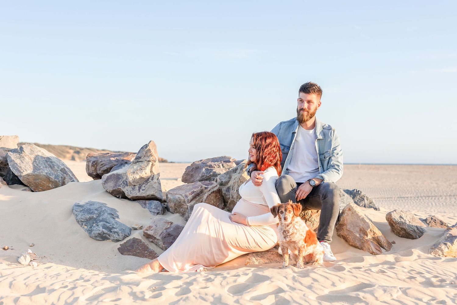 Couple with dog sitting on rocky beach during sunset maternity session.
