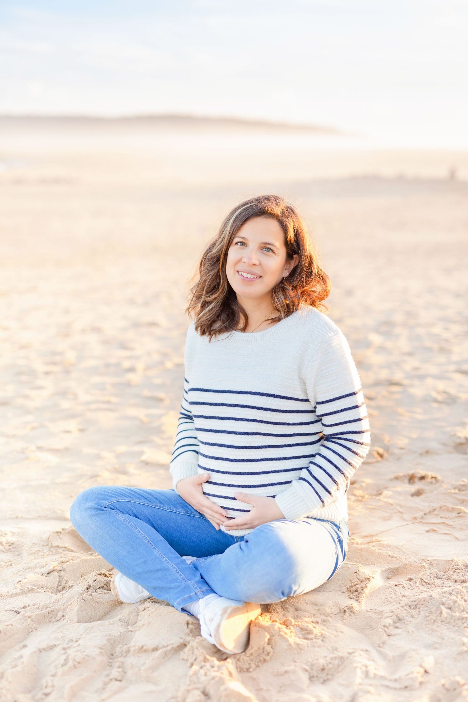 Person in striped sweater and jeans posing on sandy beach at sunset.