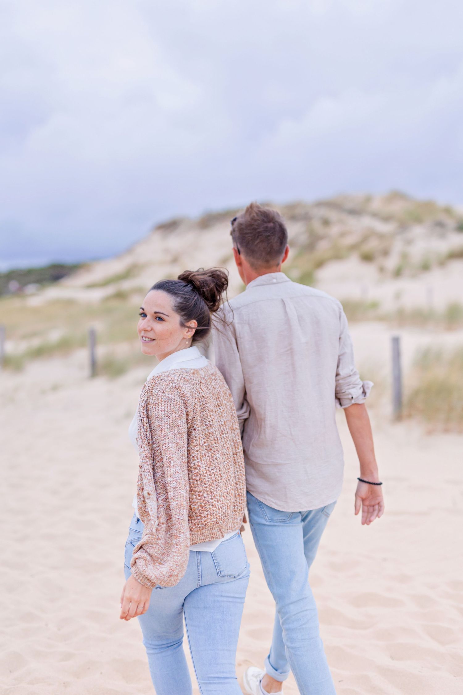 Two people strolling together on a serene beach with sand dunes and cloudy skies.