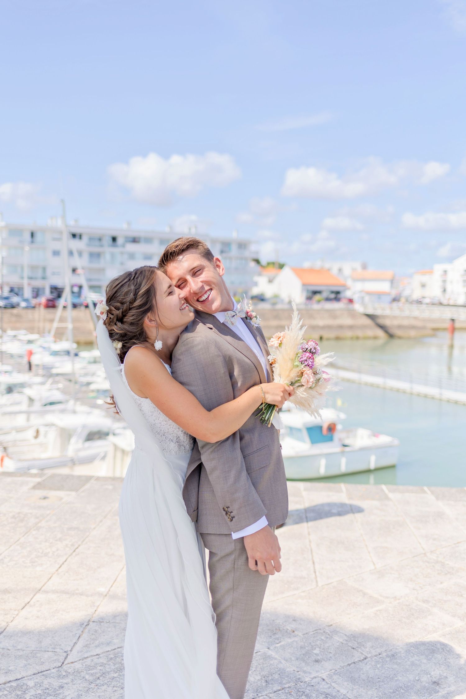 Playful wedding moment captured near marina with boats and buildings in background.