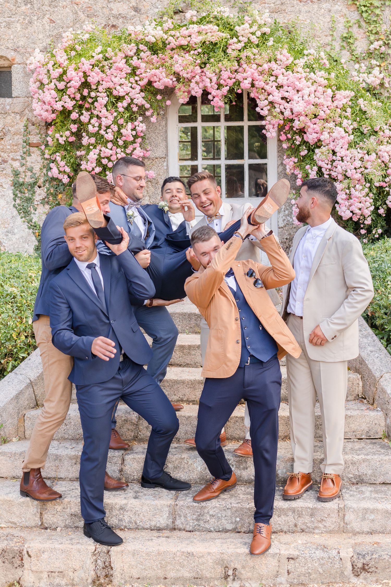 Groomsmen pose playfully on stone steps under pink flowering arch during wedding celebration.