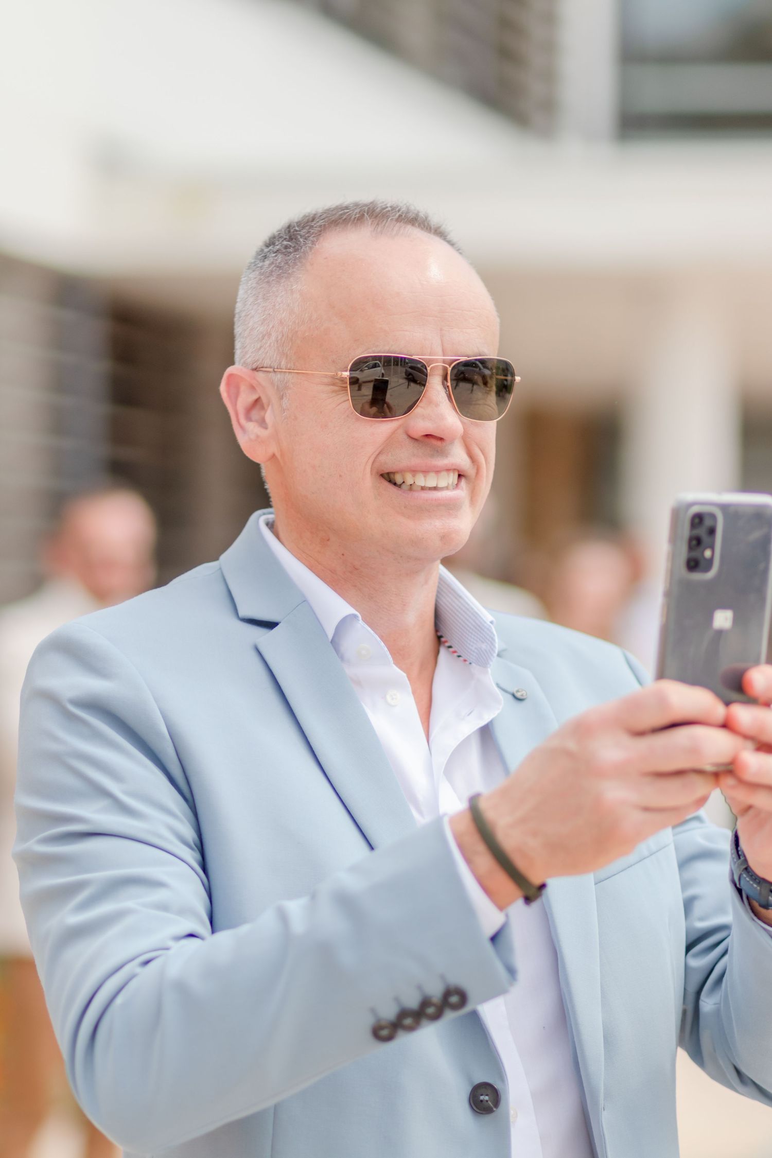Person in light blue blazer taking photos with a smartphone at an outdoor event.