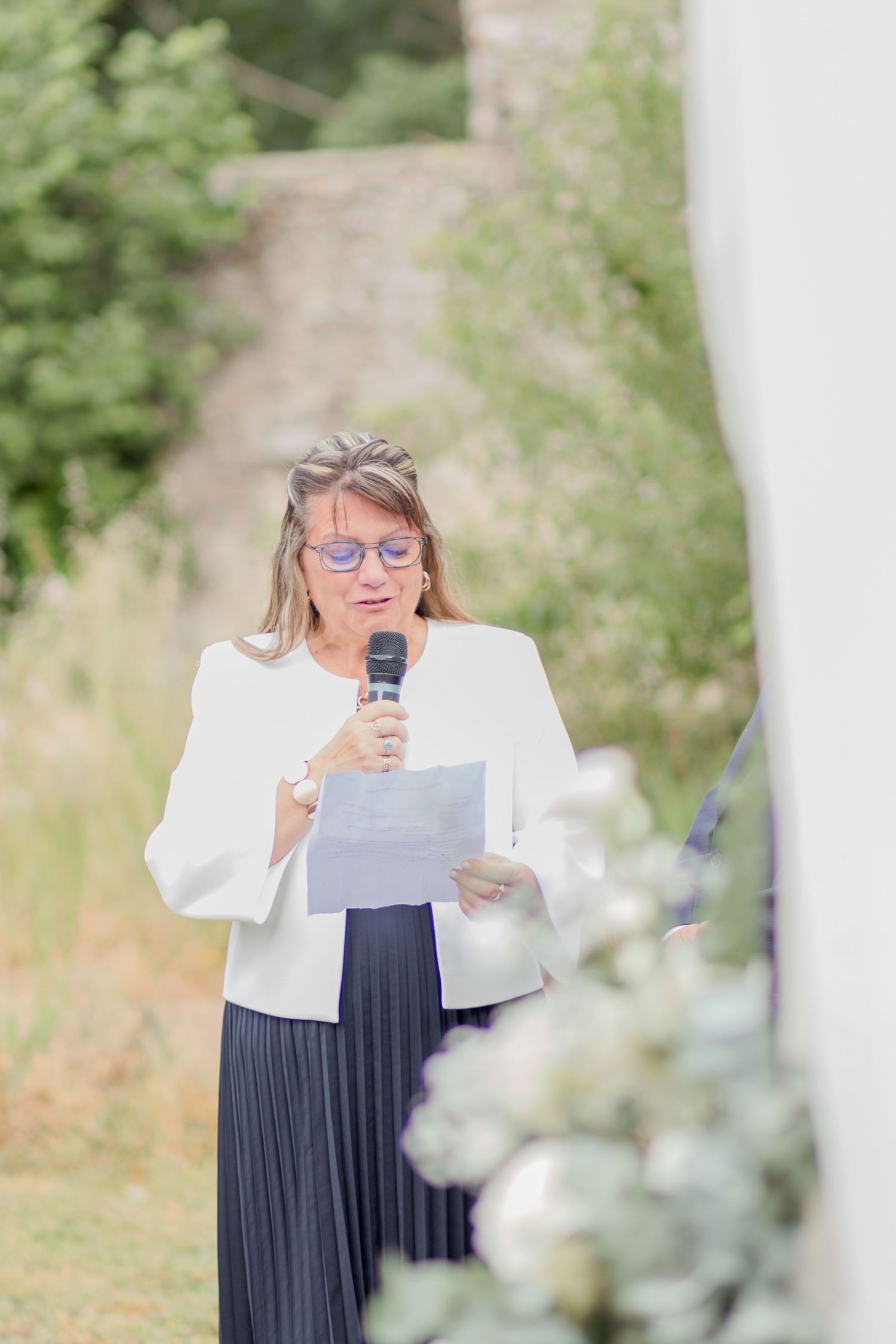 Woman in white jacket and navy dress gives speech at outdoor wedding.